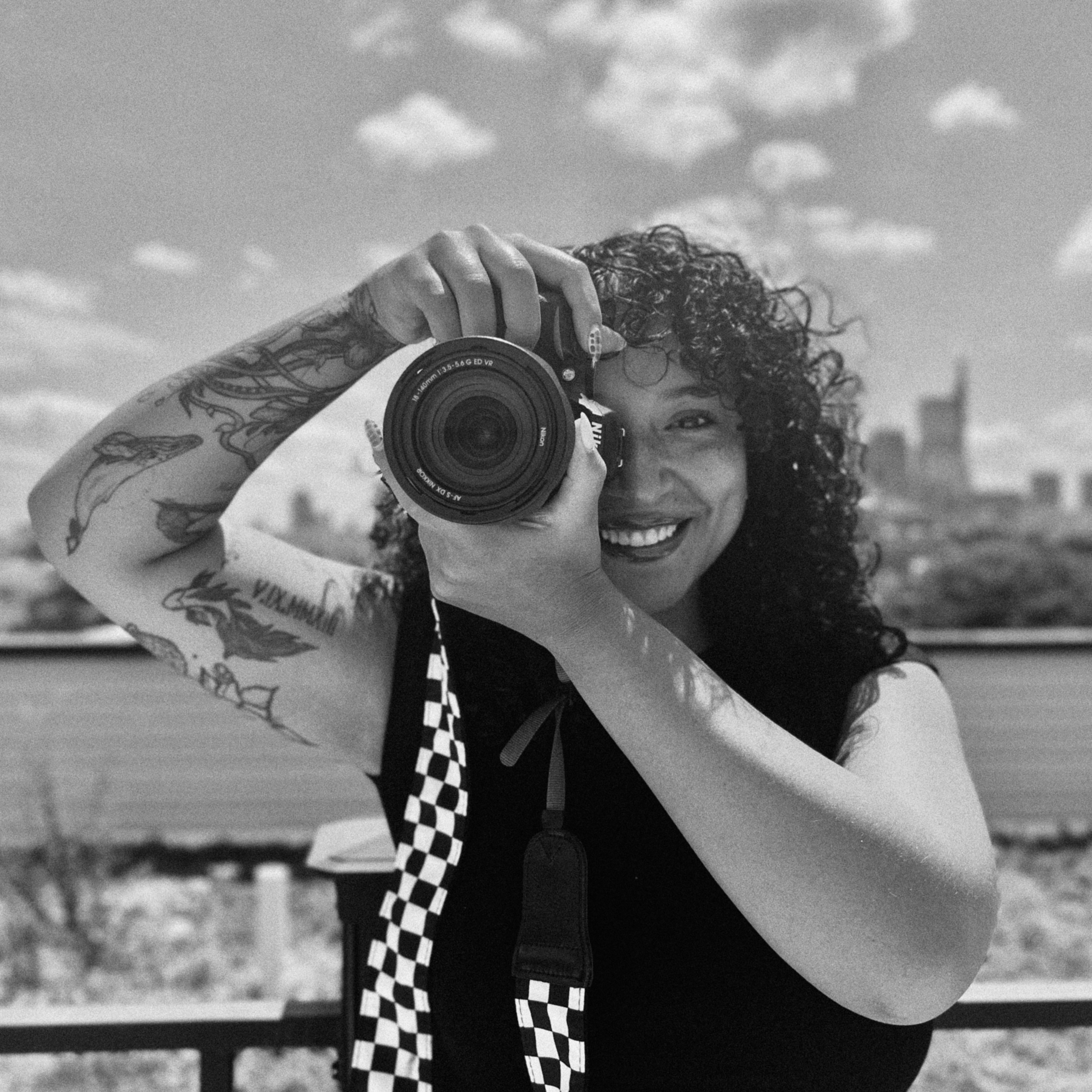 Mel holding her camera up to her eye, black and white portrait with the Philadelphia skyline behind