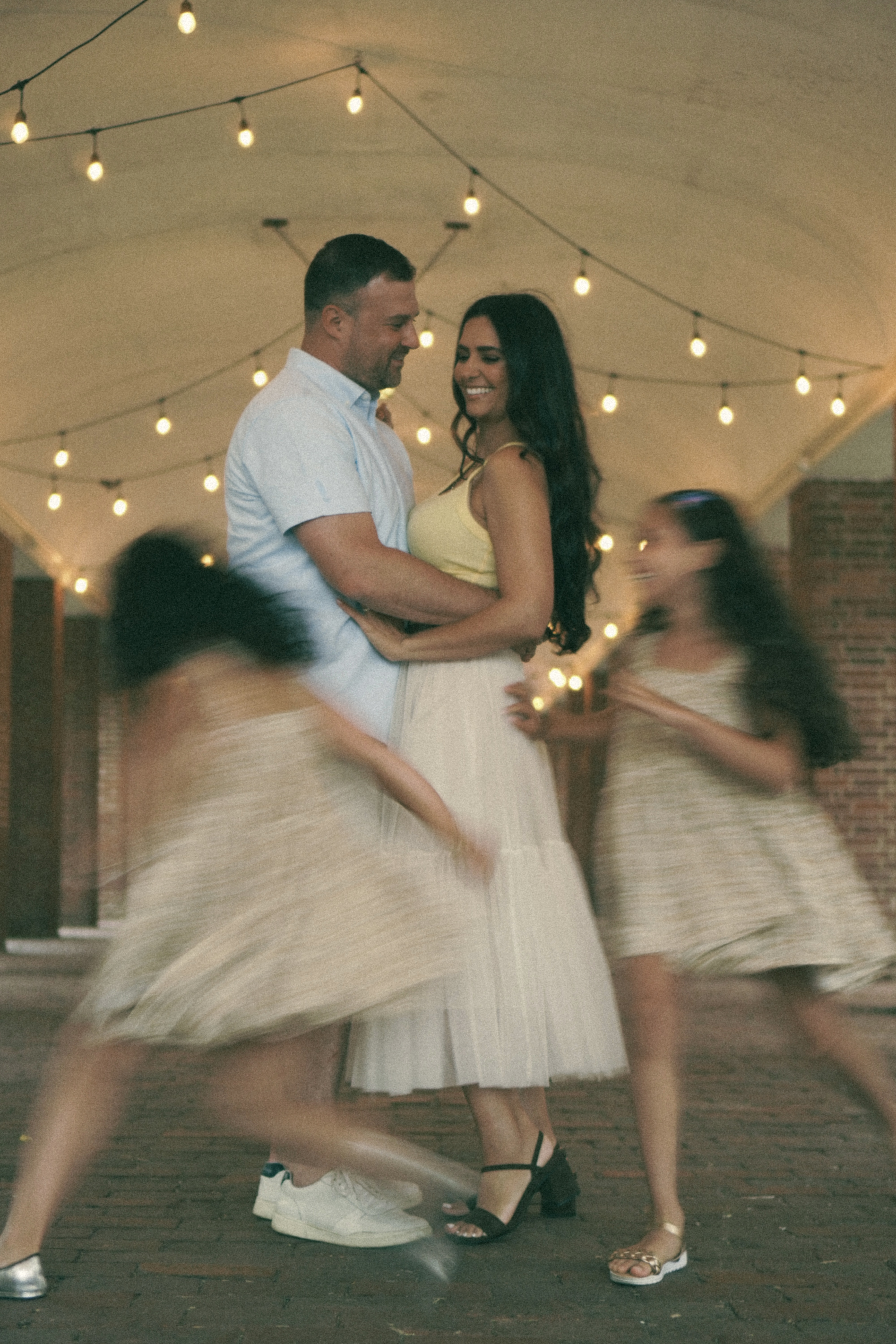 Parents slow dancing under string lights while their daughters spin around them