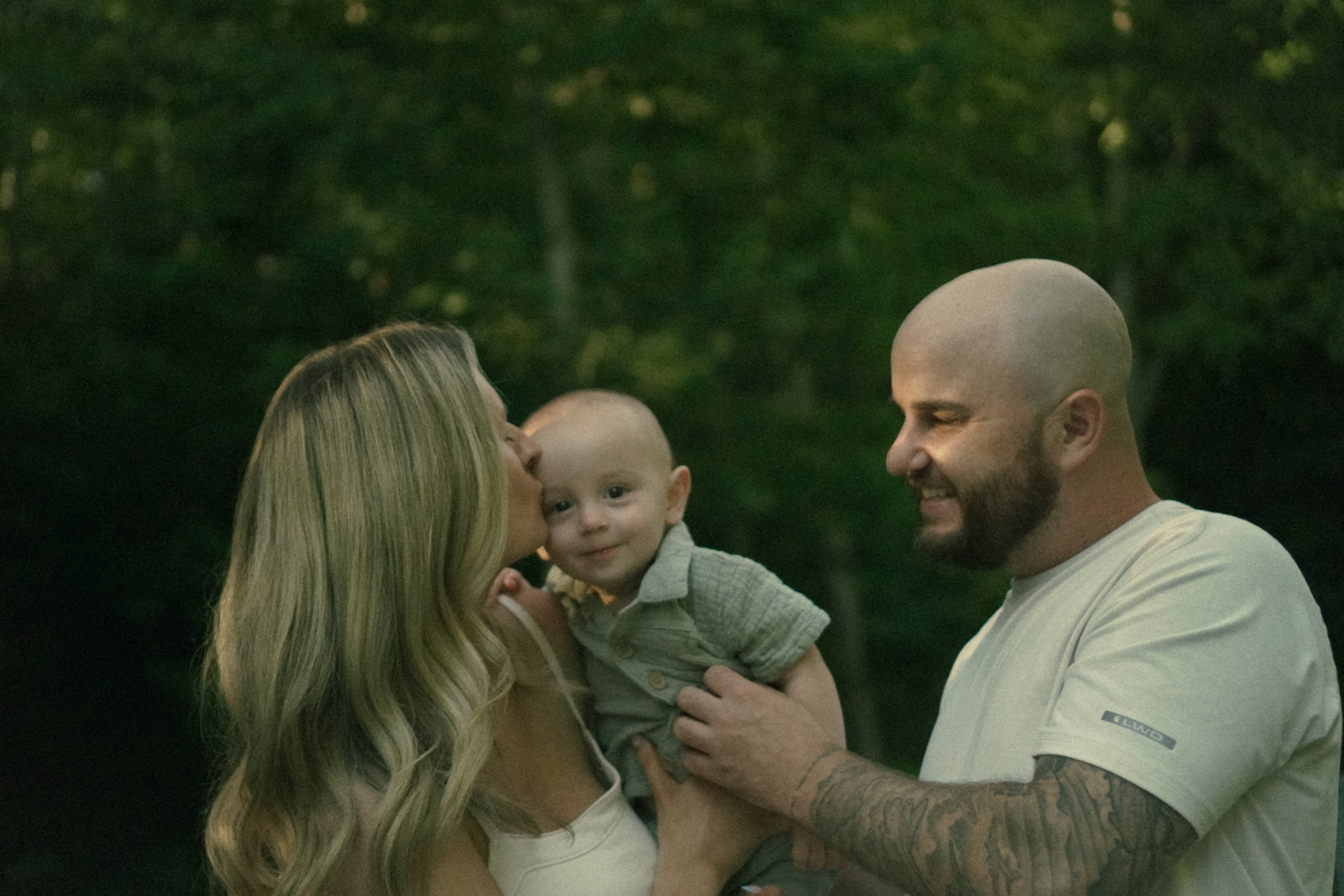 Mom kissing her baby on the cheek while dad smiles, evening light in the woods